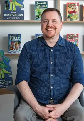 John Patrick Green, New York Times bestselling author and illustrator, sitting in front of his InvestiGators graphic novels
